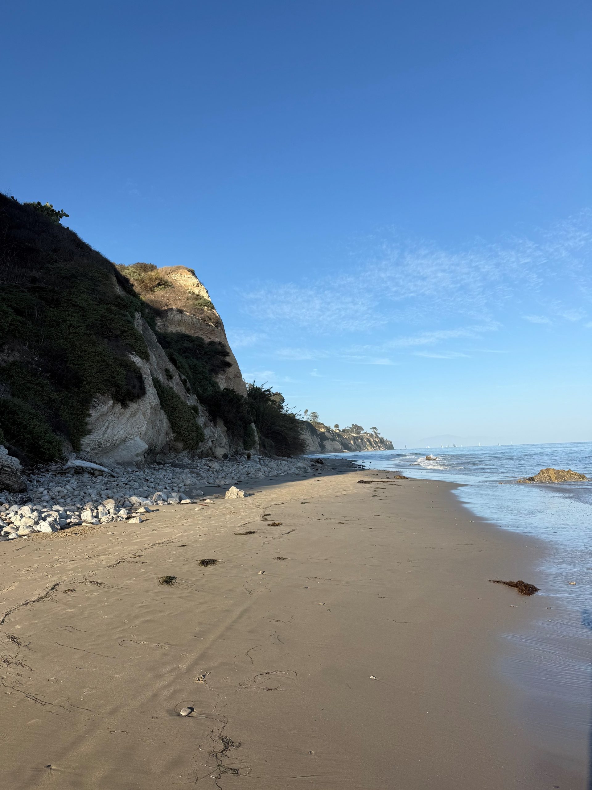 Sandy beach with a rocky cliff on the left, calm sea, and a clear blue sky with distant sailboats on the horizon.