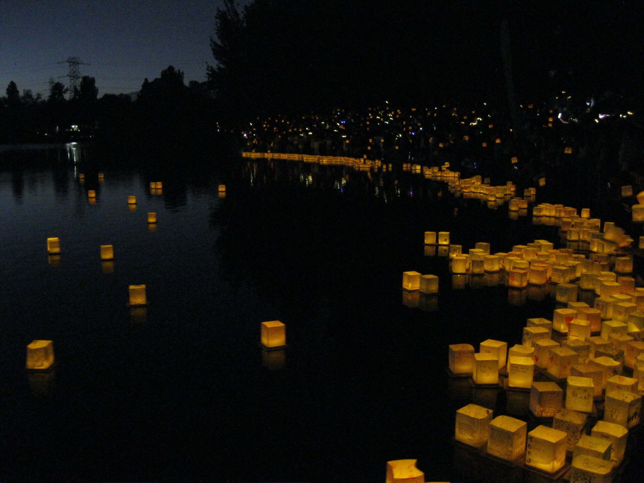 Night scene of a river at dusk filled with glowing paper lanterns floating on the water and along the shore.