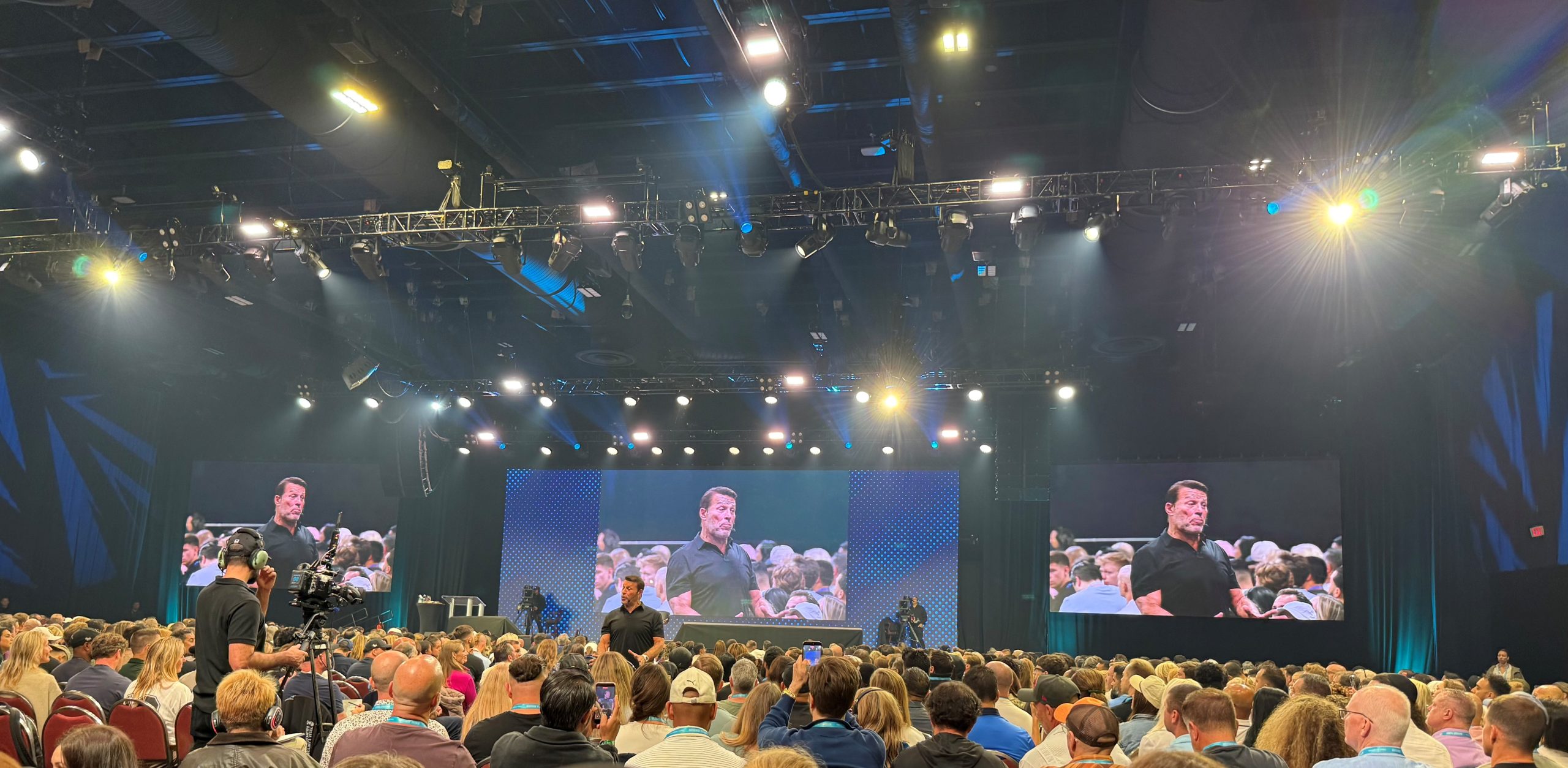 Large indoor conference hall with a stage, bright lights, and three large screens displaying a man speaking to a crowded audience