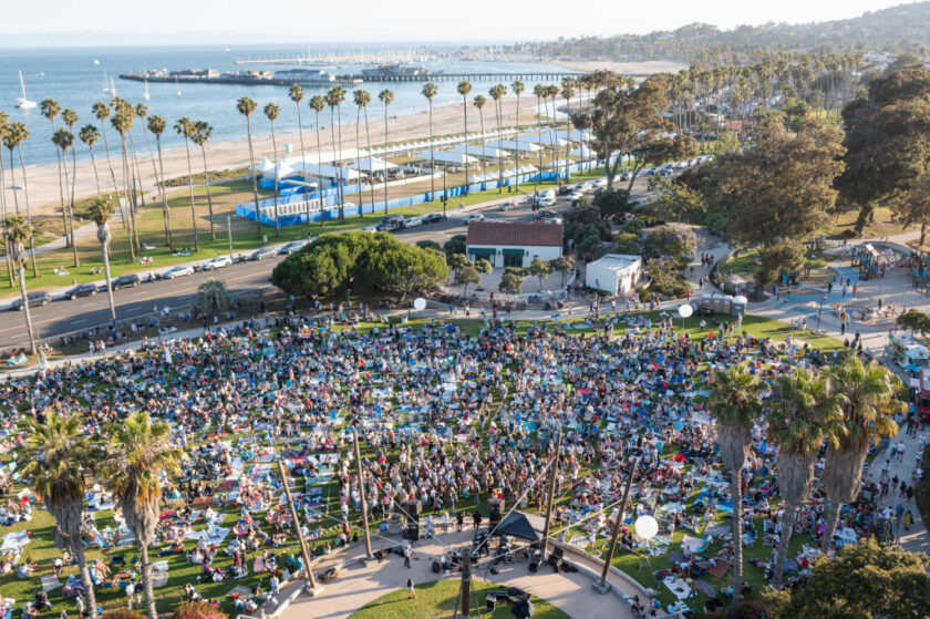 Large outdoor concert in a park by the beach, with a dense crowd seated on chairs and blankets around a central stage or performance area.