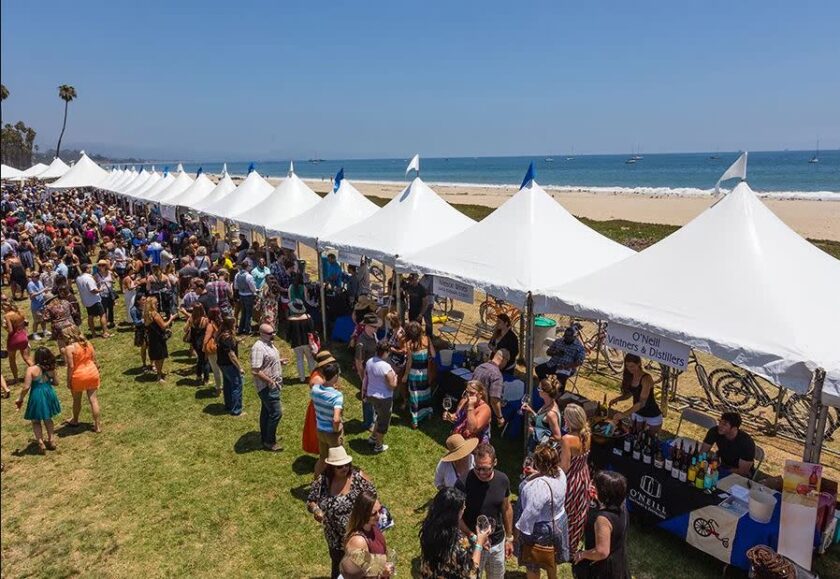 Row of white market tents set up along a beach with a crowded festival, vendors and attendees browsing stalls by the shoreline.