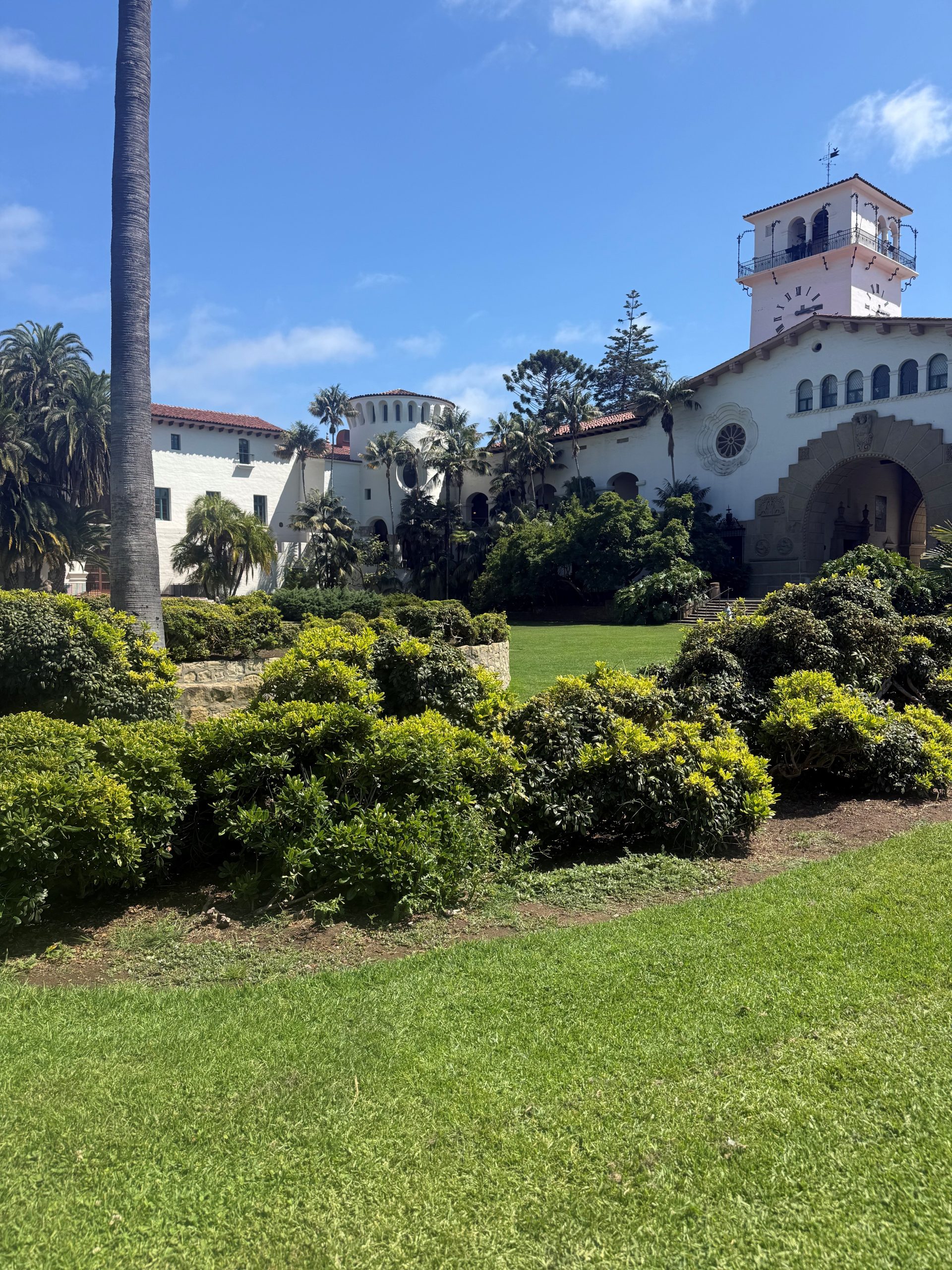 White Mediterranean-style building with a clock tower surrounded by palm trees and manicured greenery under a blue sky